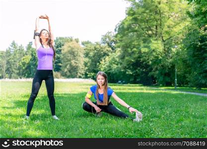 Healthy lifestyle concept - two young and fit girls stretching before jogging on the grass in a park on a sunny morning.