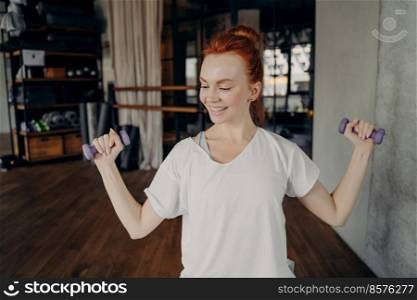 Healthy lifestyle. Young happy slim beautiful woman with red hair in bun working out in fitness studio, holding dumbbells and doing exercise for strong arms, looking away from camera and smiling. Smiling sportive redhead woman exercising with dumbbells in fitness studio