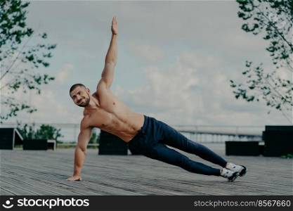 Healthy man stands in abs side plank and raises arm, does exercises outdoor, has muscular body and pleased expression. Handsome athlete trains in open air. Male couch balances on hand. Sport concept