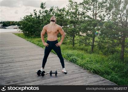 Healthy muscular man stands with naked torso, raises head above and breathes deeply, wears sport clothes, has workout with barbells, poses outdoor. Bodybuilding, active lifestyle, fitness concept