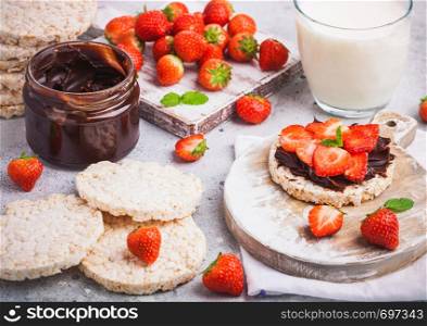 Healthy organic rice cakes with chocolate butter and fresh strawberries on wooden board and glass of milk on light stone kitchen background.