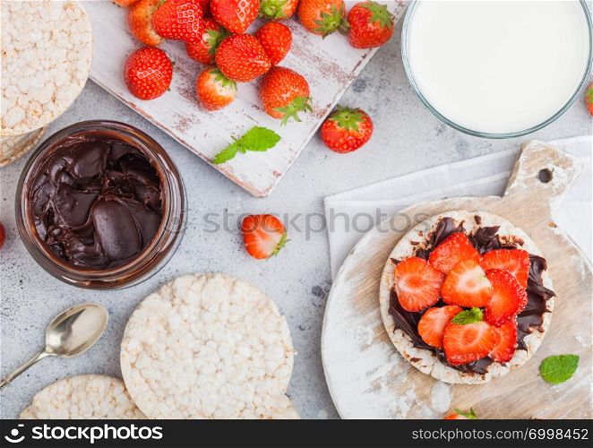 Healthy organic rice cakes with chocolate butter and fresh strawberries on wooden board and glass of milk on light stone kitchen background.