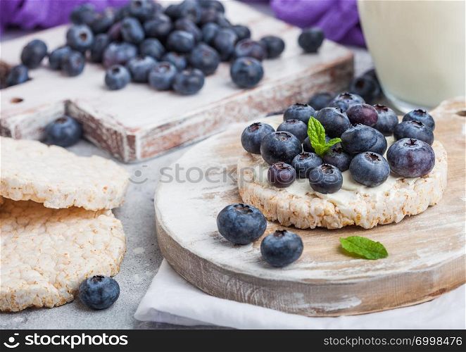Healthy organic rice cakes with ricotta and fresh blueberries on wooden board and glass of milk on light stone kitchen background.