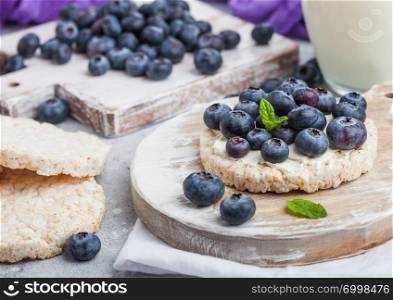 Healthy organic rice cakes with ricotta and fresh blueberries on wooden board and glass of milk on light stone kitchen background.