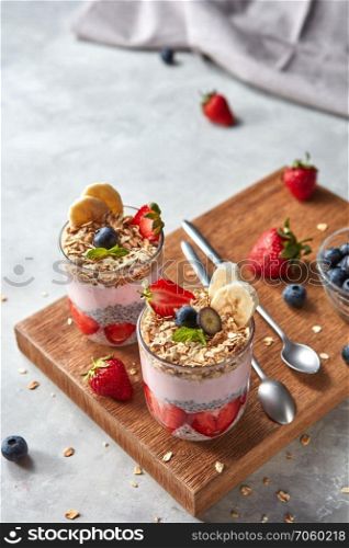 Healthy pink smoothie in glasses with natural fruits: strawberry, banana, blueberry, oat flakes and chia seeds on wooden board on grey background. Superfoods, natural detox, diet and healthy food.. Organic yogurt smoothie with strawberries, banana, blueberry, oat flakes and chia seeds, fruit dessert on wooden board on grey background