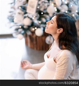 Healthy pregnant woman doing yoga exercise at home in Christmastime, with closed eyes sitting in lotus pose and meditating, inner peace and harmony