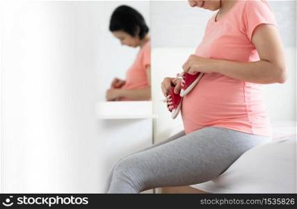 Healthy Pregnant Woman in pink shirt smiling holding Little Baby Red Shoes on Belly. Happy Asian Mother relaxing sitting on bed during her pregnancy in bedroom.