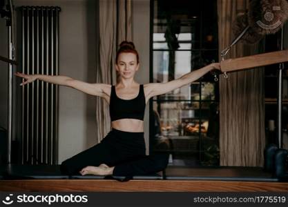 Healthy redhead female sitting on trapeze table with arms outstretched to both sides in relaxed position and smiling at camera, young woman pilates instructor doing stretching exercises in studio. Pilates instructor sitting on trapeze table with arms outstreched to both sides