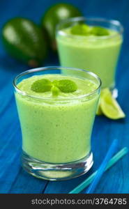 Healthy refreshing green smoothie made of fresh avocado, lime and yoghurt garnished with mint and served in glasses on blue wood (Selective Focus, Focus on the big mint leaves on the drink)