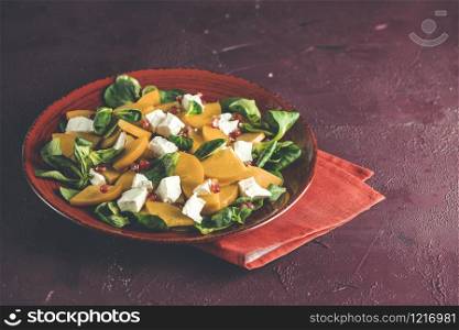 Healthy salad with persimmon, doucette (lambs-lettuce, cornsalad, feld salad) and feta cheese on a red plate on a red background. Superfoods Vitamin autumn or winter persimmon salad.