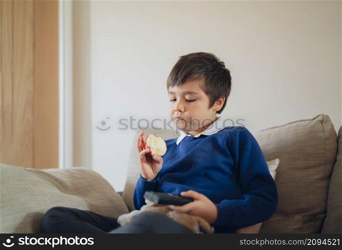 Healthy School kid eating red apple while watching TV, Portrait child eating fresh fruit for breakfast. Happy boy sitting on sofa relaxing befor go to school