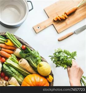 Healthy seasonal vegetables with cooking pot , cutting board and knife on white desk background. Female hand holding fresh bunch of kitchen herbs. Clean eating. Farm organic vegetables. Top view