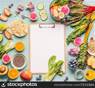 Healthy smoothie ingredients around  clipboard with blank paper sheet on light table, top view, frame. Various fruits , vegetables and berries with almond, chia seeds and pine nuts. Vegan food