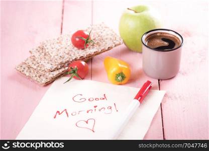 Healthy snack with whole-wheat crispbread, vegetables, apple, a cup of coffee and a napkin with a good morning message on a pink background, in daylight.