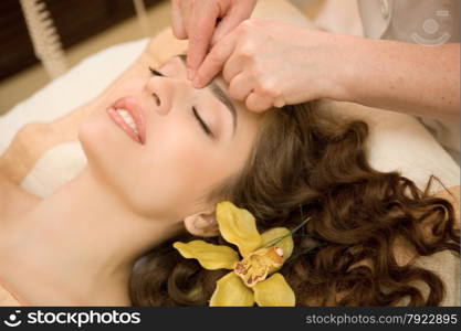 Healthy spa: young beautiful relaxing woman having a facial massage, with orchid in long brown hai