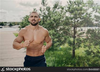 Healthy strong bearded man runner has jogging exercise in open air, breathes deeply and listens music in earphones, has naked torso, strong muscular arms. Morning workout and active lifestyle concept