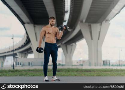 Healthy strong bodybuilder lifts heavy barbells, has workout outdoor, demonstrates his strength, has biceps, stands under bridge maintains good physical shape. Young sportsman works on muscles