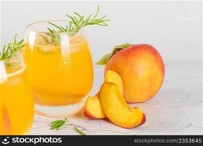 Healthy summer juice - peach cocktail with ice, rosemary twig, sugar rim, fruit slices in misted glass in elegant white interior on white marble countertop. Closeup.