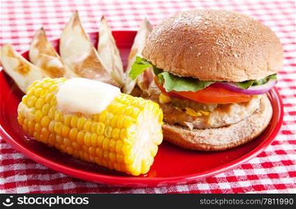 Healthy turkey burger on a whole grain bun, with baked potato wedges and corn on the cob, served on a red and white checkered tablecloth.
