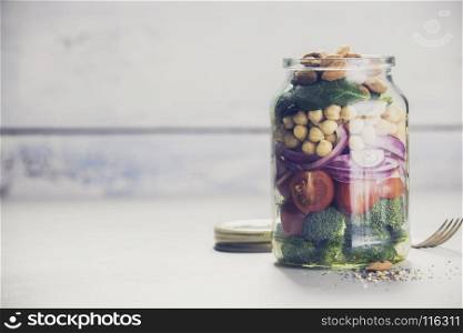 Healthy vegan energy boosting salad with chickpeas, broccoli, tomatoes, red onion, spinach and nuts in glass jar, concrete background, selective focus. Clean eating, superfood, vegan, detox food concept