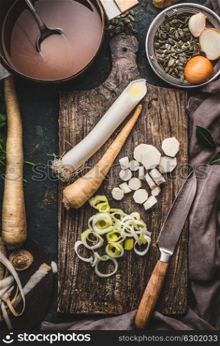 Healthy vegetables cooking and eating concept. Leek and parsnip root vegetables on cutting board with knife and pot with broth on dark rustic background, top view.