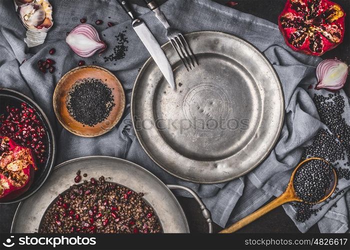 Healthy vegetarian black lentil salad with pomegranate on kitchen table with empty plate and cutlery , top view. Country dark style