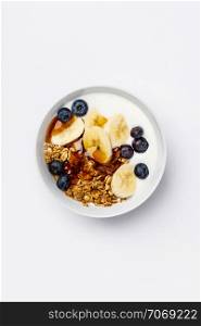 Healthy vegetarian breakfast- Oat granola with fresh blueberries, banana, soy yogurt and maple syrup on white background, flat lay, top view