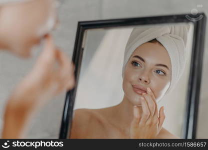 Healthy woman wears minimal makeup, takes care of complexion and lips, looks at herself in mirror, stands bare shoulders, wears bath towel on head, has healthy flawless skin. Beauty, hygiene concept