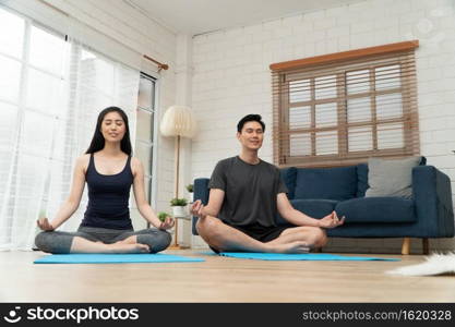 Healthy young Asian couple attractive couple practicing yoga exercises sitting on a mat in lotus pose in an online class on quarantine at home. Concept of Technology for New normal lifestyle