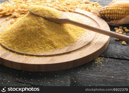 Heap of corn flour on a wooden platter with a wooden spoon on top of it, surrounded by corn cob, grains and cereal flakes, on a black wooden background.