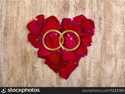 Heart made of red petals with golden wedding rings on wooden table