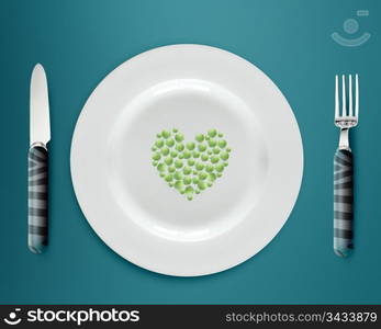 Heart shape green peas on white plate with knife and fork on blue background.