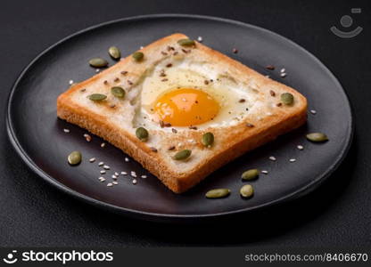 Heart shaped fried egg in bread toast with sesame seeds, flax seeds and pumpkin seeds on a black plate on a dark concrete background