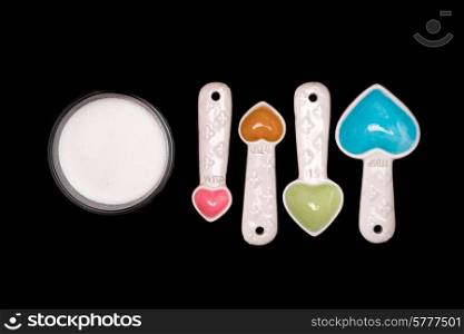 Heart shaped measuring spoons next to a bowl of sugar on an isolated black background.