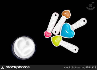 Heart shaped measuring spoons next to a small bowl of sugar with a hard shape inside the sugar, all on an isolated black background.