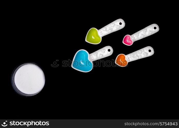 Heart shaped measuring spoons of diffferent sizes and colors facing the direction of a round bowl of sugar bringing together the ideas of love, conception and food, or sweet stuff. All on a black isolated background.