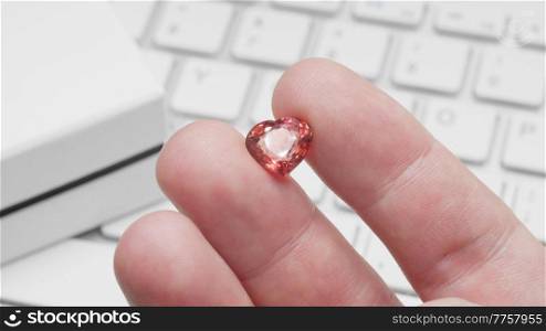 Heart shaped tourmaline gem in hand of gemologist or jeweller. Online jewelry shopping for birthday, Valentine Day or other holiday. Male hand holding red tourmaline, macro shot with box and keyboard.. Online jewelry shopping with heart shaped tourmaline