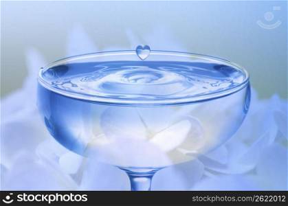 Heart shaped water droplet falling into a vlear glass with flowers in the background