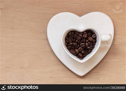 Heart shaped white plate and cup filled with roasted coffee beans on wooden table background. Top view