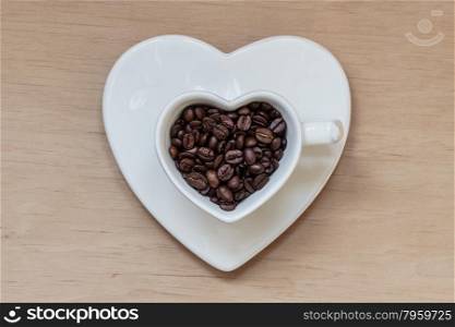 Heart shaped white plate and cup filled with roasted coffee beans on wooden table background. Top view