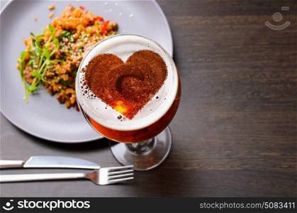 Heart silhouette on foam in beer glass on black table with food, view from above.