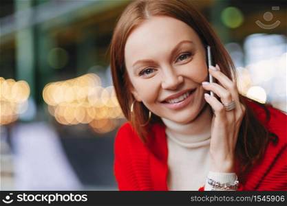 Heashot of lovely European female entrepreneuer has healthy skin, pleasant smile, dressed in red clothes, looks straight at camera, holds modern cell phone, poses outside, blurred background