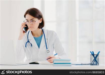 Heathcare personnel, medicine concept. Serious brunette female doctor focused at modern laptop computer, rewrites necessary information, talks on mobile phone, calls someone, has serious look