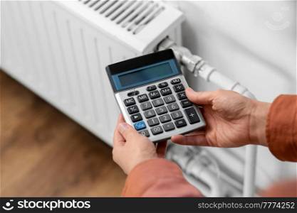 heating, energy crisis and consumption concept - close up of hand with calculator and radiator at home. close up of hand with calculator and home radiator