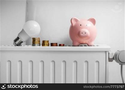 heating, energy crisis and consumption concept - piggy bank, light bulb and money on radiator at home. piggy bank, light bulb and coins on radiator