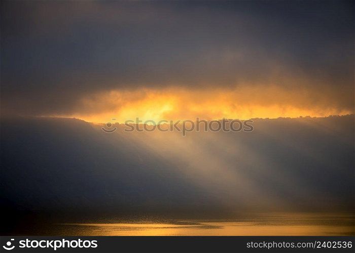 Heavy clouds over the rocky shore. Sunrays barely break through, illuminate small houses and the water surface. Sunbeams Through Heavy Clouds