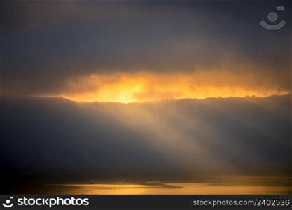 Heavy clouds over the rocky shore. Sunrays barely break through, illuminate small houses and the water surface. Sunbeams Through Heavy Clouds