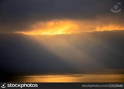 Heavy clouds over the rocky shore. The sun?s rays barely break through, illuminate small houses and the water surface. Background with Sunbeams Through Heavy Clouds
