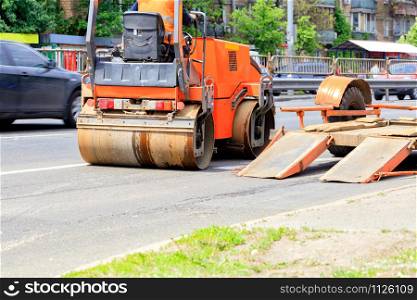 Heavy vibrating rink is unloaded from a low platform trailer and rides along the road on asphalt pavement on a city street, image with copy space.. Heavy vibrating road roller is unloaded from a low platform trailer and stands on the roadway.