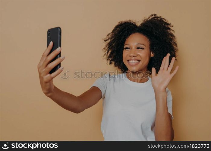 Hello! Beautiful cheerful dark skinned woman smiling at camera on modern smartphone, taking selfie on mobile phone and feeling happy, gesturing hi while making video call, isolated over studio wall. Cheerful african woman gesturing hi while making video call or taking selfie on smartphone